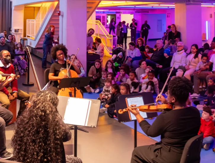 D-Composed playing in front of kids sitting on floor mats and an audience seated in the Harris Theater lobby.