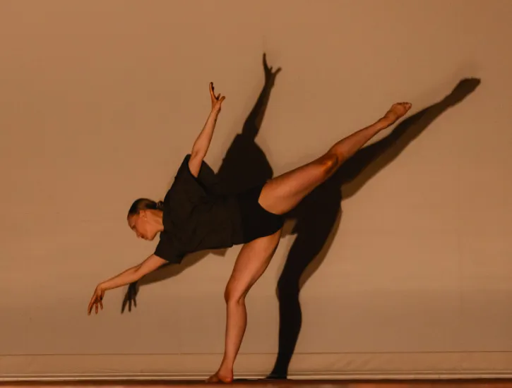 A dancer reaches to the ground gracefully while extending her leg up. The light casts a shadow on the wall behind her.