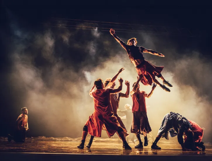 A dancer jumping in the air and four other dancers catching him. Fog covers them all.