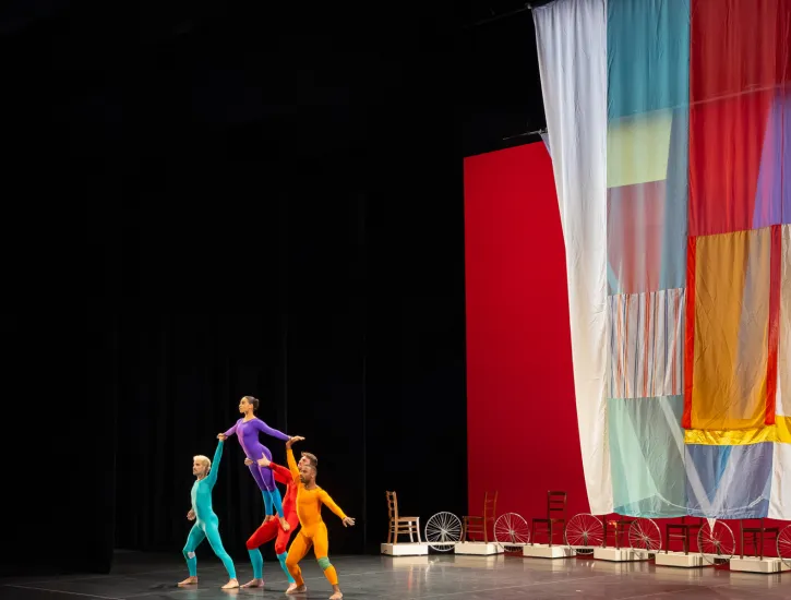 Four dancers in colorful body suite hold up one dancer as she leans forward. A colorful drape flows behind them. A row of chairs also display behind the dancers.