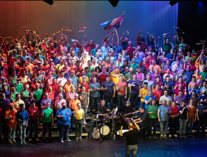 A men's chorus on stage singing. Everyone is wearing different color shirts.