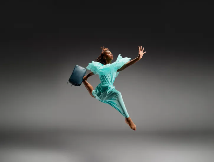 A young dancer jumping in the air wearing a blue dress and carrying a suit case.