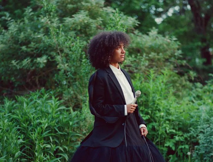 A woman in a black and white gown holding a microphone in the middle of the woods.