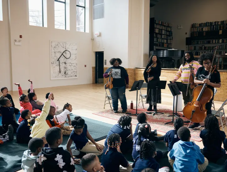 D-Composed String Ensemble interacting with children who are sitting on floor mats.